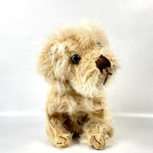 May include: A plush, seated dog toy with a shaggy, light brown fur. The toy has dark brown eyes and a darker brown nose. The dog's paws are also a darker brown color. The toy is sitting upright, facing to the right.