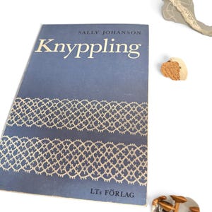 May include: A blue softcover book titled Knyppling by Sally Johanson, featuring white lace-patterned graphics on the cover and the text LT's F&Ouml;RLAG at the bottom. The book is positioned next to small, textured, tan-colored craft items.