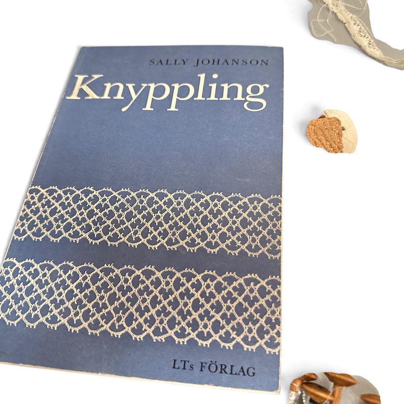 May include: A blue softcover book titled Knyppling by Sally Johanson, featuring white lace-patterned graphics on the cover and the text LT's F&Ouml;RLAG at the bottom. The book is positioned next to small, textured, tan-colored craft items.
