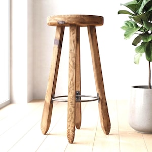 May include: A tall wooden stool featuring a round seat and three angled legs made of natural brown wood with visible grain patterns. A circular metal ring connects the three legs near the base, providing structural support for the stool.