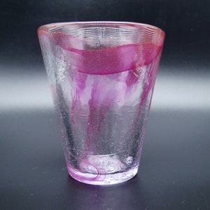 Puede incluir: Un vaso de vidrio transparente con forma acampanada. El borde y el interior presentan un color magenta vibrante, con vetas del mismo color que caen por los lados. El vidrio tiene un aspecto texturizado.