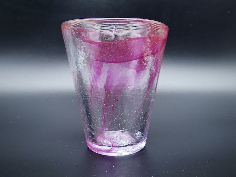 Puede incluir: Un vaso de vidrio transparente con forma acampanada. El borde y el interior presentan un color magenta vibrante, con vetas del mismo color que caen por los lados. El vidrio tiene un aspecto texturizado.