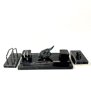 May include: A black marble desk set featuring a rectangular base with a bronze-toned dog figurine, a letter opener, two square containers, a lidded box, and a metal letter holder. Each piece displays white veining throughout the polished stone.
