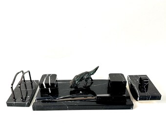 May include: A black marble desk set featuring a rectangular base with a bronze-toned dog figurine, a letter opener, two square containers, a lidded box, and a metal letter holder. Each piece displays white veining throughout the polished stone.