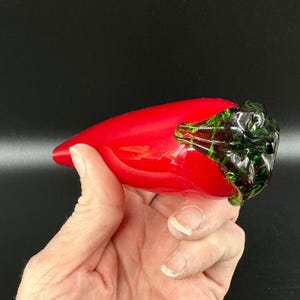 May include: A smooth, glossy red glass chili pepper figurine featuring a dark green glass stem at the top. The object is shaped like a tapered chili pepper with a rounded end and a textured, multi-faceted green glass cap at the base of the stem.