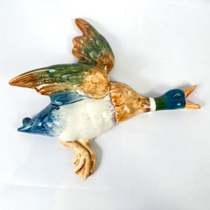 May include: A ceramic wall-mounted mallard duck figurine in flight, featuring a glossy finish with a dark green head, a white neck ring, brown and green wings, a white chest, and a blue tail. The duck has an open beak and extended feet.