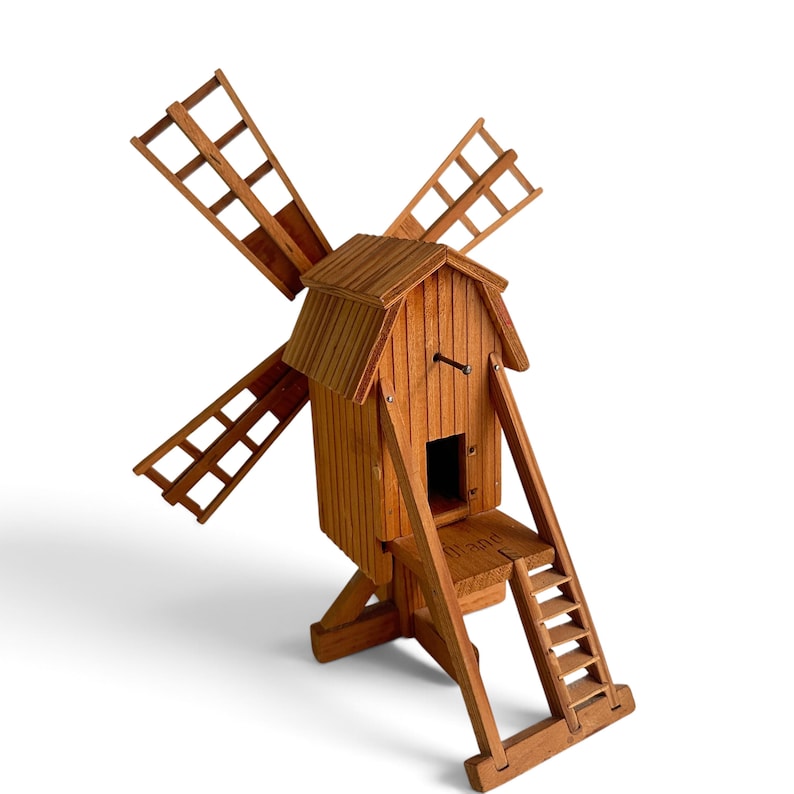 May include: A wooden windmill model featuring a brown finish, four lattice-style blades, a peaked roof, and a small attached ladder leading to a platform. The word Holland is clearly engraved on the wooden platform base of the structure.