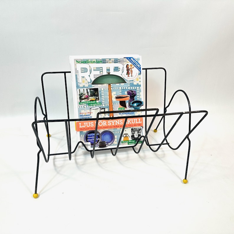 May include: A black metal magazine rack with a mid-century modern design. The rack holds a magazine with the text "Scandinavian Retro" and "Ljus or Syns Kull". The rack has yellow feet.