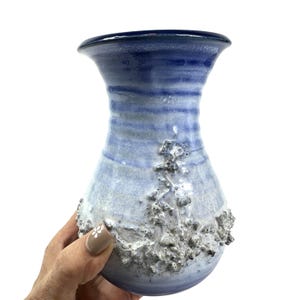 May include: A ceramic vase featuring a flared rim and a tapered neck. The body displays a gradient of light blue to deep blue hues with horizontal bands. A prominent, textured, white, craggy, and raised surface detail adorns the lower section.
