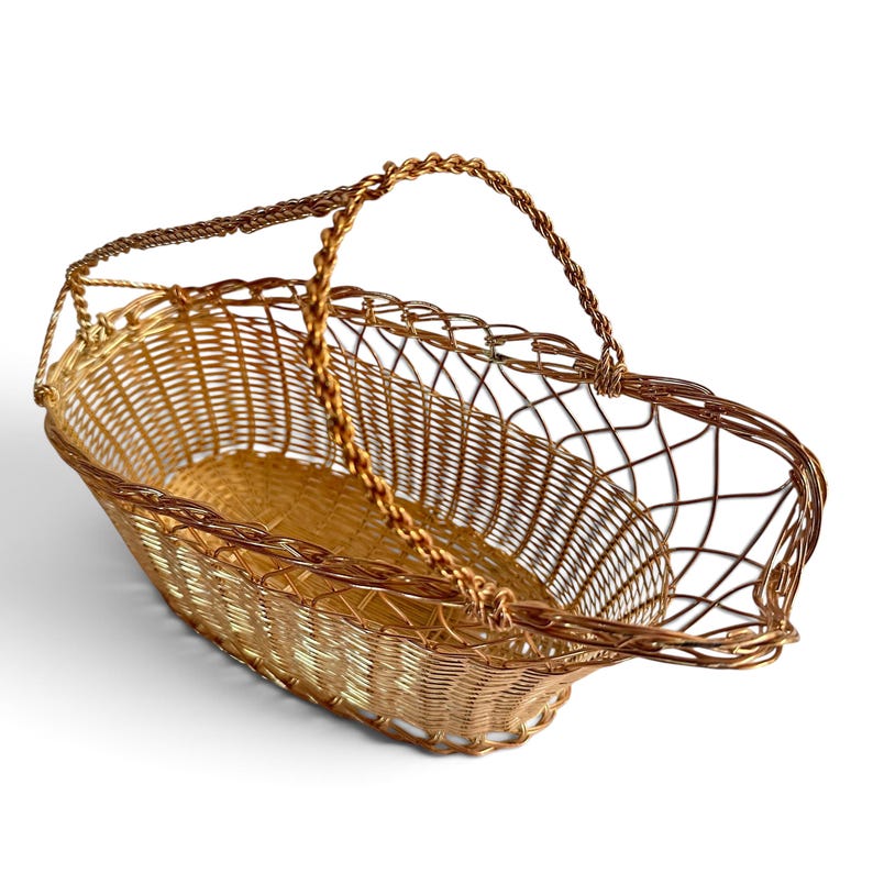 Puede incluir: Una cesta de forma ovalada construida con alambre met&aacute;lico fino de tono dorado. La cesta presenta una base y laterales de malla tejida fina, con un borde decorativo de alambre trenzado y una sola asa arqueada de alambre trenzado.