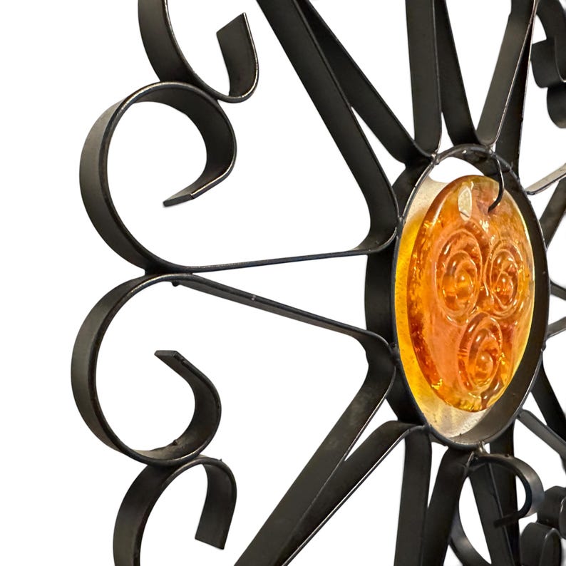 Peut inclure: Une d&eacute;coration murale en m&eacute;tal noir pr&eacute;sentant un motif en forme de soleil avec des p&eacute;tales incurv&eacute;s en volutes rayonnant autour d'un accent central en verre ambr&eacute; circulaire orn&eacute; de trois motifs en spirale en relief.