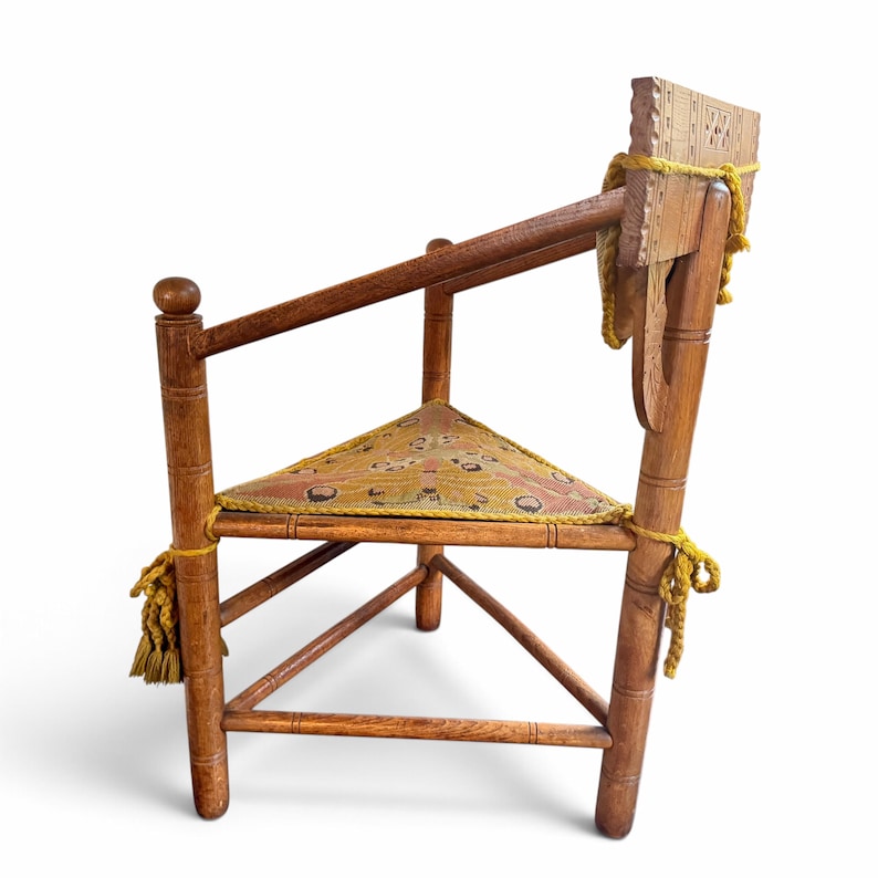 May include: A wooden corner chair with a triangular seat featuring a yellow and brown patterned fabric cushion. The chair has turned legs, a carved backrest, and yellow decorative tassels tied to the frame at the seat corners.