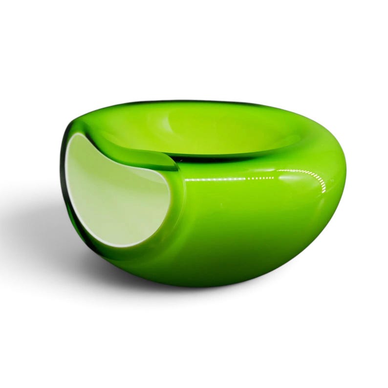 May include: A smooth, rounded glass bowl featuring a vibrant lime green exterior and a contrasting white interior. The bowl has a wide, open top and a distinct, curved indentation on one side, creating a unique, asymmetrical, and organic shape.