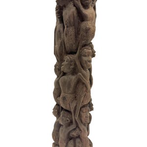 May include: A tall, dark brown wooden sculpture featuring a vertical column of multiple human figures carved in relief. The figures are stacked on top of one another, with detailed facial features, arms, and torsos visible throughout the structure.