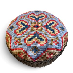 May include: A round pincushion featuring a cross-stitch pattern on a light blue background. The design includes geometric shapes in navy blue, red, and tan thread. The base of the cushion is finished with a dark brown, gathered fabric trim.