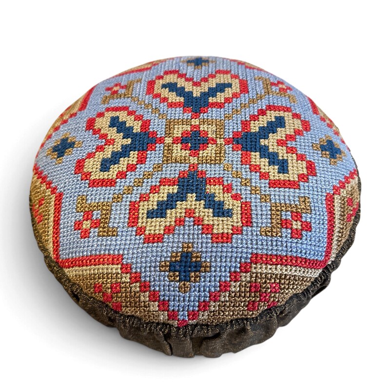 May include: A round pincushion featuring a cross-stitch pattern on a light blue background. The design includes geometric shapes in navy blue, red, and tan thread. The base of the cushion is finished with a dark brown, gathered fabric trim.