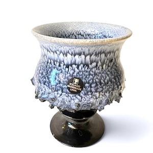 Puede incluir: Un cáliz de cerámica con borde acampanado y base de pedestal. La parte superior presenta un esmalte texturizado y moteado en tonos blanco, gris y negro. Una pegatina negra circular muestra el texto ICELAND HANDMADE LAVA en blanco.