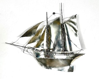 May include: A wall-mounted metal sculpture of a sailing ship crafted from polished silver-toned sheet metal. The vessel features multiple masts, thin wire rigging, and curved sails that appear to be catching the wind, creating a dynamic effect.