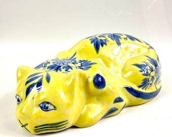 May include: A ceramic cat figurine in a reclining position, featuring a bright yellow glazed finish with hand-painted blue floral and leaf patterns across its back, head, and tail. The cat has blue painted eyes and whiskers on its face.