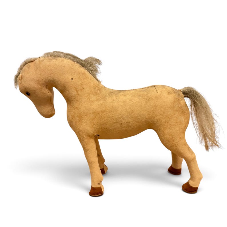 May include: A tan fabric horse toy with a soft, felt-like texture, featuring a light-colored yarn mane and tail, dark brown stitched hooves, and small black bead eyes. The horse is depicted in a standing position with its head tilted downward.