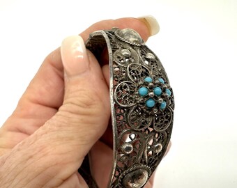 May include: A silver-toned hinged cuff bracelet featuring intricate filigree metalwork and a central floral motif. The flower is accented with small, round, bright turquoise-colored stones arranged in a cluster at the center of the petals.