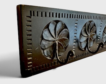 May include: A rectangular black wooden wall rack featuring three carved circular floral motifs. Each flower center serves as a base for a curved black metal hook. The wooden board is decorated with a carved border of repeating vertical rectangular notches.