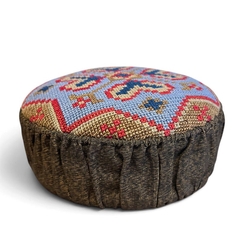 May include: A round, flat cushion featuring a top surface with a cross-stitch pattern in light blue, tan, red, and dark blue. The sides of the cushion are made of a dark, textured fabric with vertical pleats gathered around the circumference.