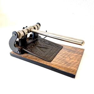 Puede incluir: Una perforadora de papel de estilo vintage con una base de metal oscuro y un diseño en relieve de la letra A. El mecanismo incluye una palanca larga de metal plateado pulido y dos cabezales de perforación cilíndricos sobre una base de madera.