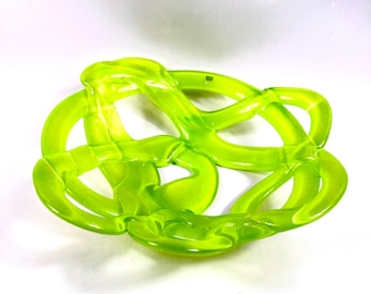 May include: A bright green, abstract decorative object with a woven, knot-like design. The translucent piece is made of a glossy material and sits on a white surface. The object's shape is reminiscent of a Celtic knot.