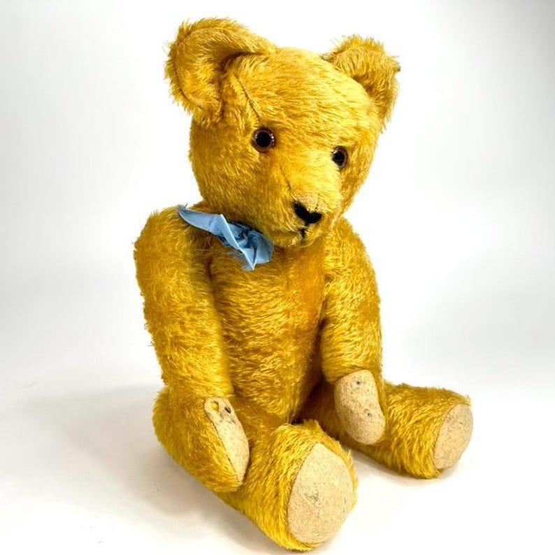 May include: A vintage golden-yellow teddy bear with a light blue ribbon around its neck. The bear has dark eyes, a black nose, and tan paw pads. It is sitting upright against a white background, showcasing its classic design.
