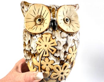 May include: A ceramic owl figurine featuring a textured cream and brown mottled glaze. The owl has large, round, light-coloured eyes with dark pupils and a black beak. The body is decorated with raised, light-coloured flower motifs.