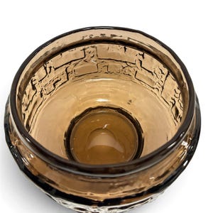 May include: A round, amber-colored glass bowl featuring a textured, raised brick-like pattern on the interior walls. The glass is translucent, with a smooth, circular base and a thick, dark-tinted rim that defines the opening of the vessel.