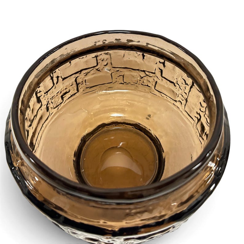 May include: A round, amber-colored glass bowl featuring a textured, raised brick-like pattern on the interior walls. The glass is translucent, with a smooth, circular base and a thick, dark-tinted rim that defines the opening of the vessel.