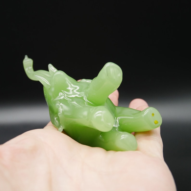 K&ouml;nnte beinhalten: Eine durchscheinende, hellgr&uuml;ne Figur eines stilisierten Tieres, m&ouml;glicherweise eine Schildkr&ouml;te oder ein &auml;hnliches Wesen, mit abgerundeten Merkmalen und gelben Akzenten. Die Figur wird in einer Hand vor dunklem Hintergrund gehalten.