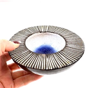 Peut inclure: Un bol en céramique fait à la main, avec un extérieur marron foncé et un intérieur blanc et bleu. Le bord présente un motif radial de lignes noires et blanches. Le bol est tenu par une main.