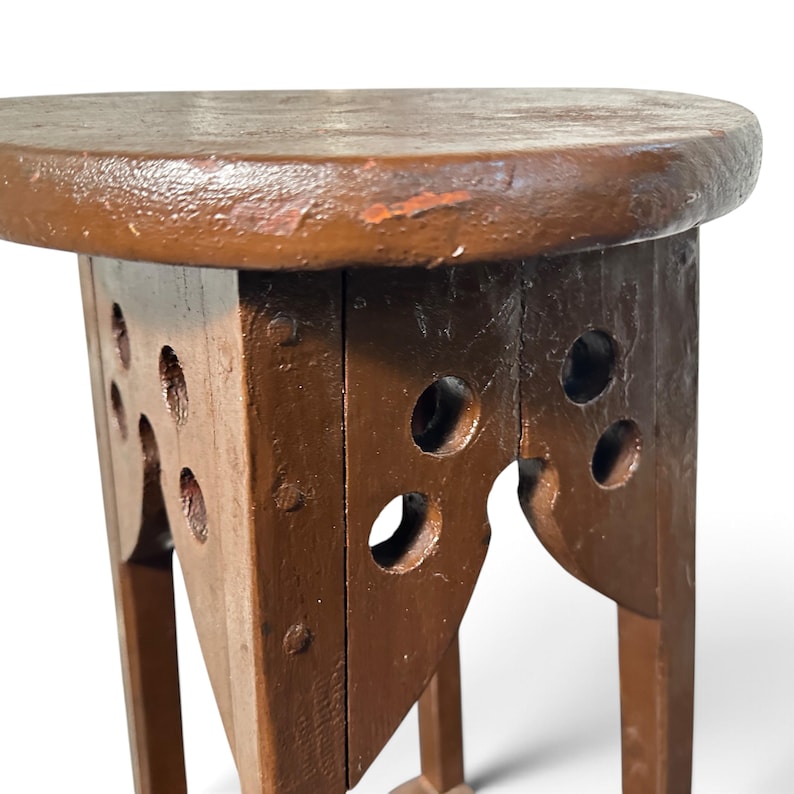 May include: A brown wooden stool with a round top and four legs. The upper section of the legs features decorative cutouts with circular holes and arched edges. The wood has a painted finish with visible signs of wear and texture.