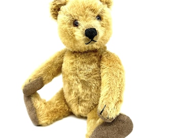 May include: A golden-tan plush teddy bear with a fuzzy, textured coat, dark brown stitched nose, and small black eyes. The bear features contrasting brown fabric on the soles of its feet and palms, and is posed in a seated position.