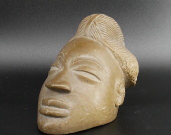May include: A tan-colored stone sculpture of a human head featuring stylized, closed eyes, arched eyebrows, a prominent nose, and full lips. The hair is depicted with detailed, linear carvings, and the ears are represented by textured circular shapes.