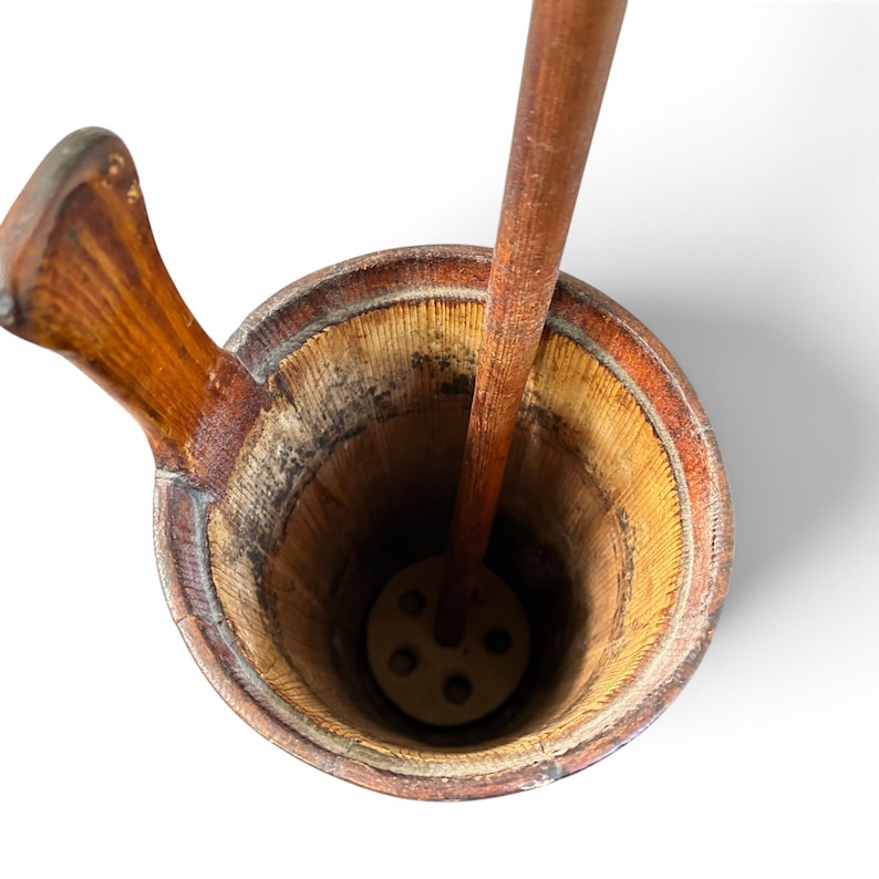 May include: A cylindrical wooden butter churn featuring a tall, vertical plunger with a perforated circular base. The interior walls show a textured, grain-heavy finish, while a single handle extends from the upper rim of the vessel.