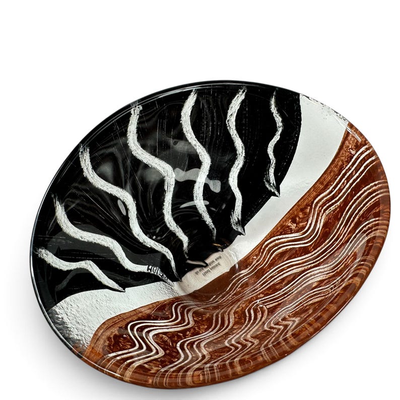 May include: A round glass bowl featuring a bold design with a black section decorated with vertical white wavy lines and a brown section featuring horizontal wavy white lines. The center of the bowl contains small printed text that reads "loving heart, new soul, one of".