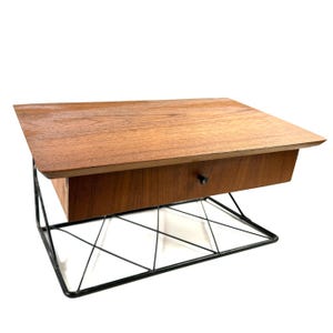 May include: A mid-century style side table with a rectangular wooden top and a single drawer. The drawer has a small, round, black knob. The table is supported by a black wireframe base.
