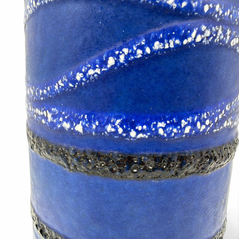 May include: A cylindrical ceramic vase featuring a deep blue matte finish. The surface is decorated with horizontal bands of textured black glaze and diagonal stripes of white speckled glaze that wrap around the body of the vessel.