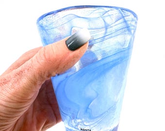 Op de afbeelding: Een lichtblauw glazen drinkglas met een wervelend, gemarmerd patroon door het doorschijnende materiaal. De onderkant van het glas is duidelijk gemarkeerd met de zwarte tekst Kosta Boda, wat de fabrikant van dit drinkglas aangeeft.