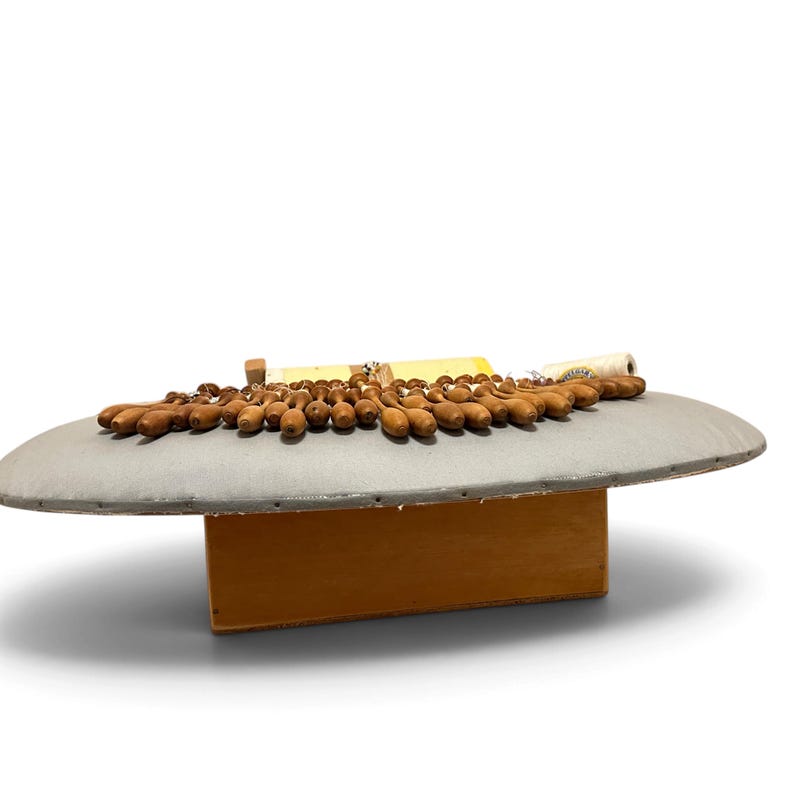 May include: A lace pillow with a gray fabric surface and a wooden base, featuring numerous brown wooden bobbins arranged in a row along the top edge, with a spool of white thread and a small paper label visible in the center.