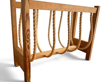 May include: A rectangular magazine rack constructed from light brown wood with a visible grain. The frame features four vertical wooden posts connected by horizontal bars, with thick, natural-colored twisted rope loops hanging between them.