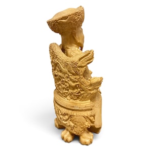 Op de afbeelding: Een lichtbeige uitgesneden beeldje van een zittende figuur met een textuurhoed en traditionele gewaden. De figuur zit op een versierde stoel met gedetailleerde reli&euml;fs van draken en wolken, rustend op vier uitgesneden poten in dierlijke vorm.