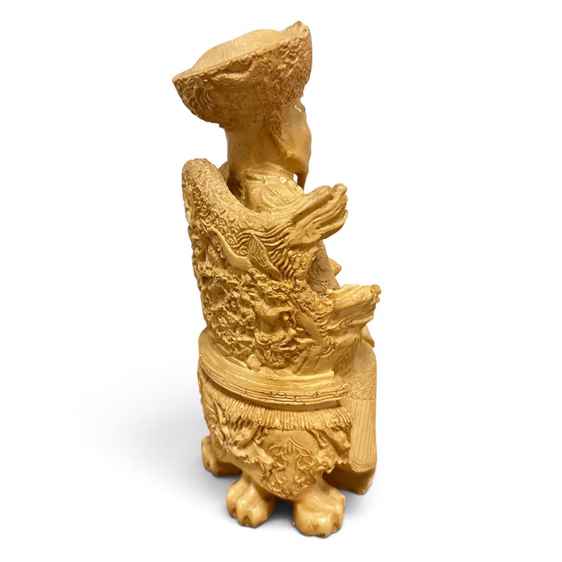 Op de afbeelding: Een lichtbeige uitgesneden beeldje van een zittende figuur met een textuurhoed en traditionele gewaden. De figuur zit op een versierde stoel met gedetailleerde reli&euml;fs van draken en wolken, rustend op vier uitgesneden poten in dierlijke vorm.