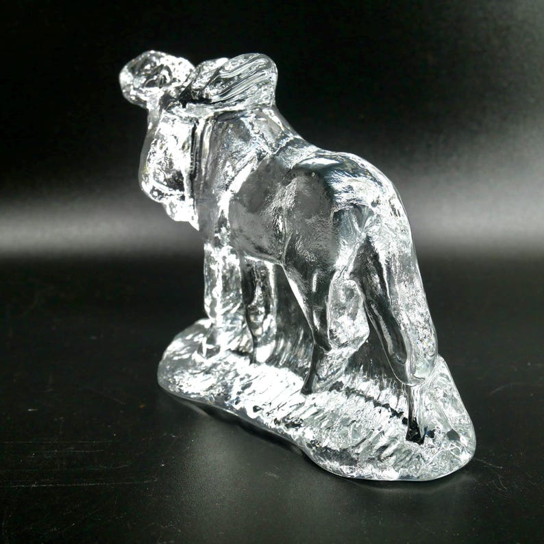 May include: A clear glass sculpture of a horse standing on a textured base. The horse is depicted in a realistic style, with details visible throughout the body. The sculpture is set against a dark background.