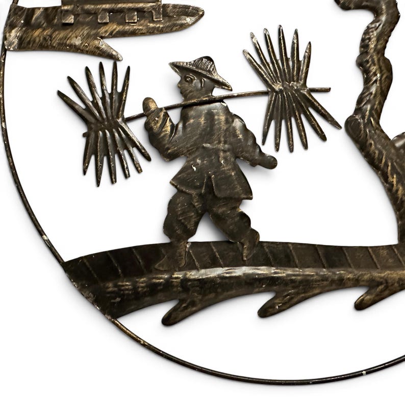 May include: A circular metal wall art piece featuring a dark bronze finish. The design depicts a figure wearing a hat, walking on a textured path while carrying a pole across their shoulders with two large, spiky bundles attached to each end.