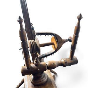 May include: A brown wooden spinning wheel featuring a central flyer assembly with a bobbin, a U-shaped flyer arm with metal hooks, and turned wooden support posts. The structure includes a horizontal base and vertical components with decorative finials.